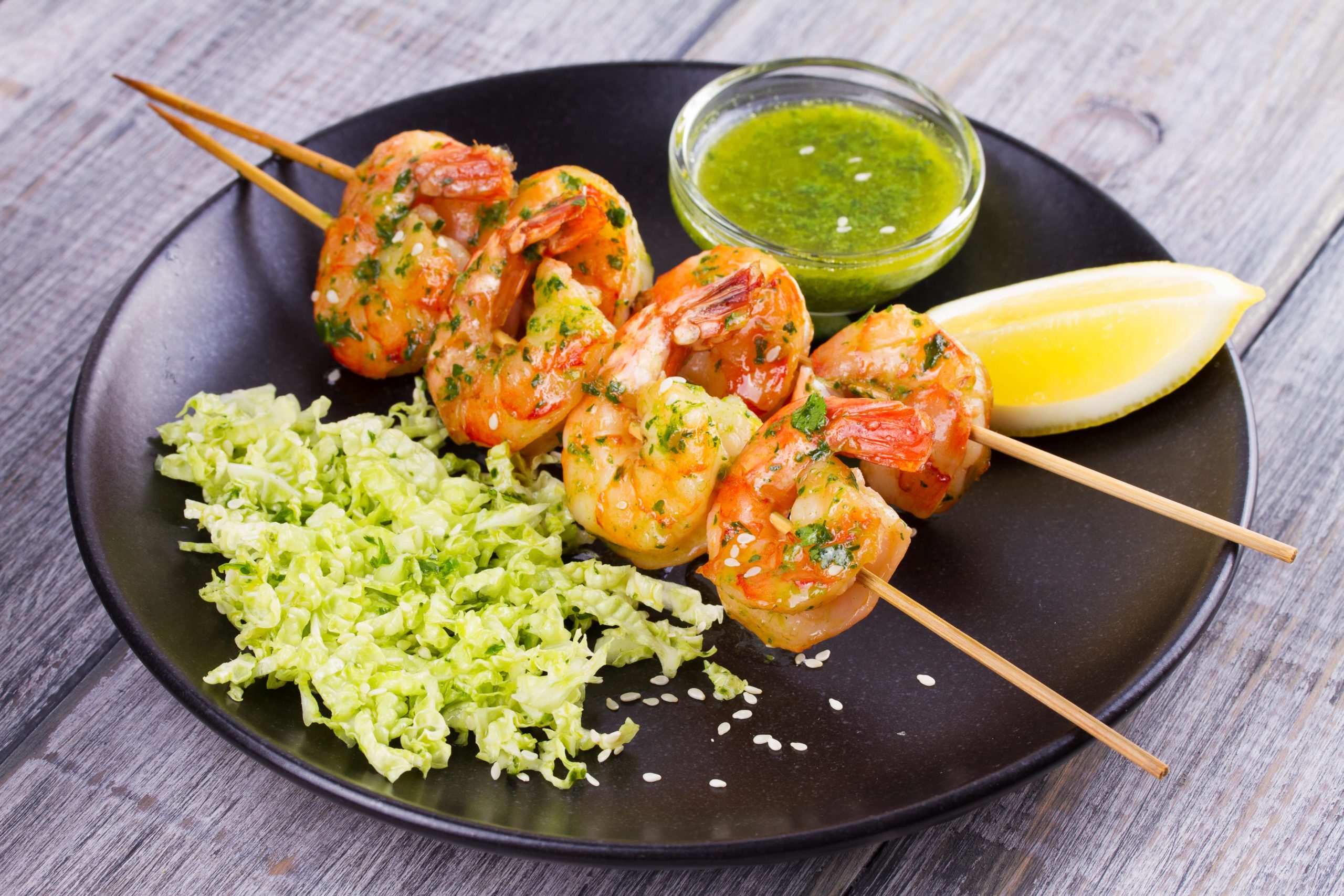 grilled shrimp on skewers alongside a lemon wedge and ramekin of pesto sauce, all served on a black plate